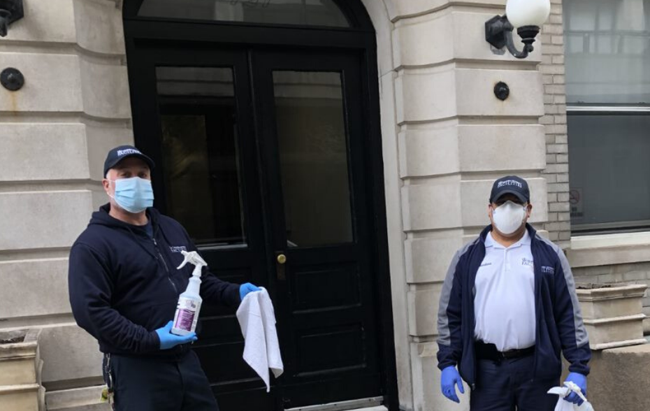 two men with wearing face masks, blue gloves and Columbia branded jacket and hat in front of residential building