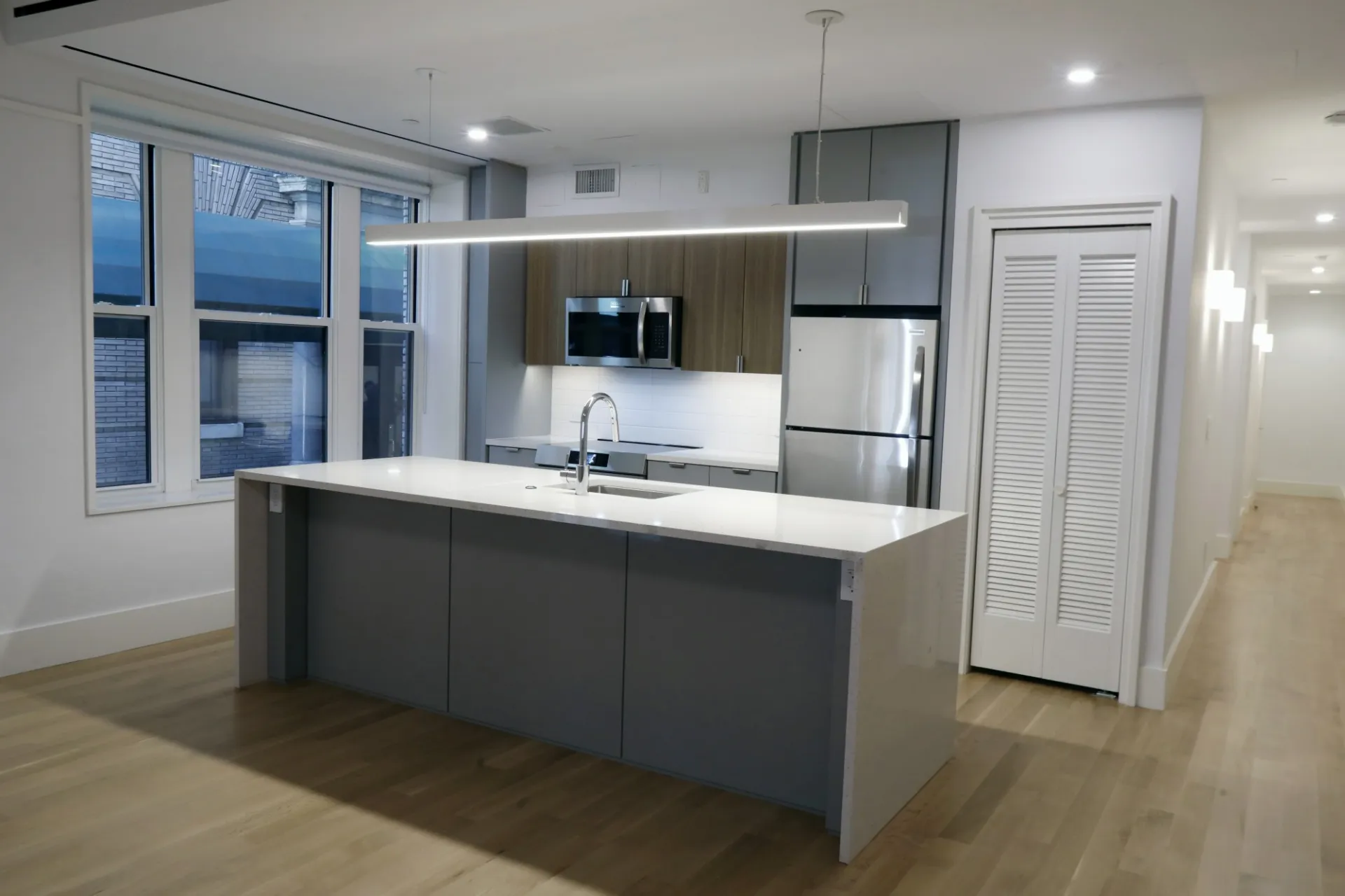 A view of the renovated kitchen which has a gray and white granite kitchen island, gray and wood accent cabinetry, and a white backsplash